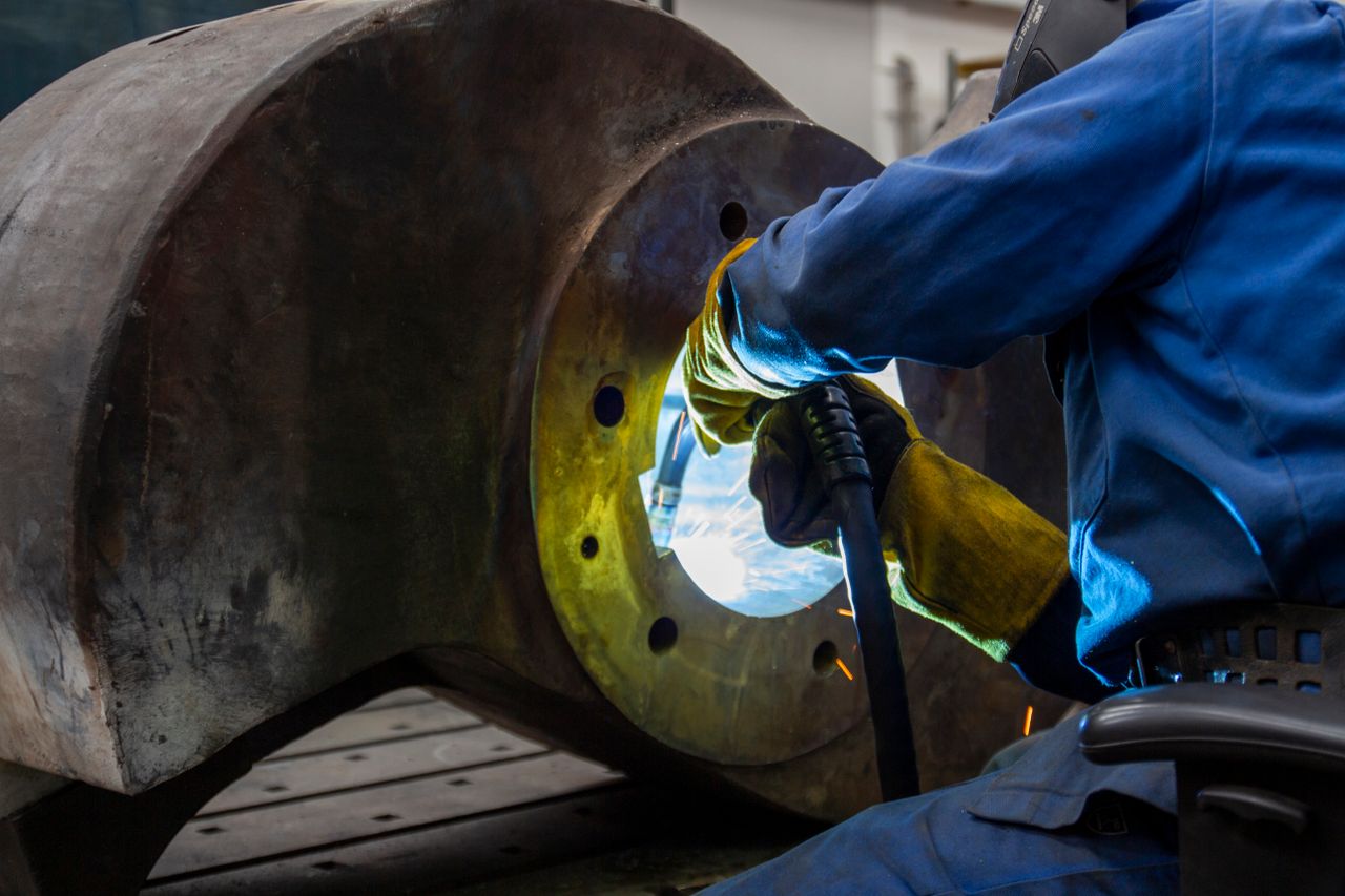 Servicios Welder operates inside a large metal machine part. Industrial setting with sparks visible.